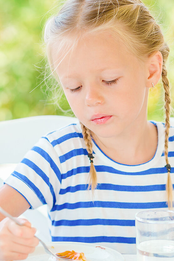 A girl in a striped shirt eating at a table, looking thoughtful.