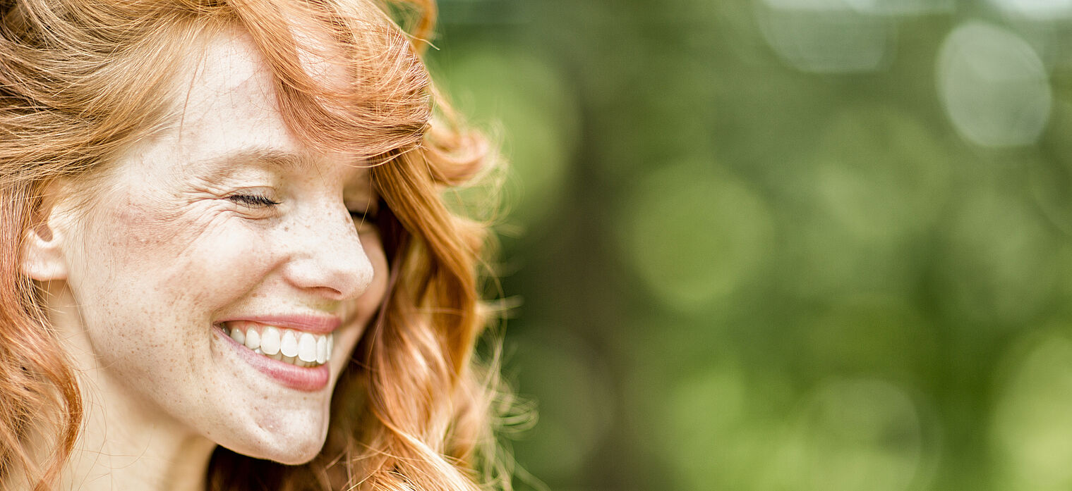 A woman with red hair smiling brightly, with a green, blurred background.