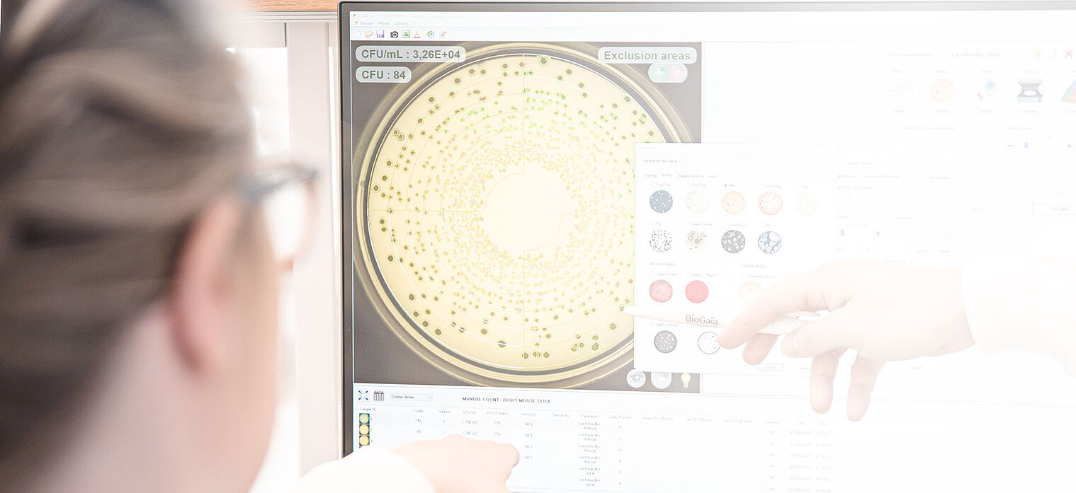 Scientist pointing at a computer screen displaying a petri dish with bacterial colonies and a color-coded chart, used for laboratory analysis.