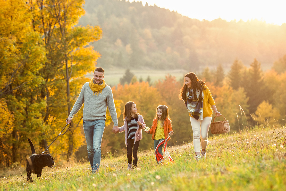 A family of four, including two young girls, walking through a sunny autumn landscape with colorful trees. The father is holding a leash of a black dog, while the mother carries a basket. They are all smiling and enjoying the outdoors.